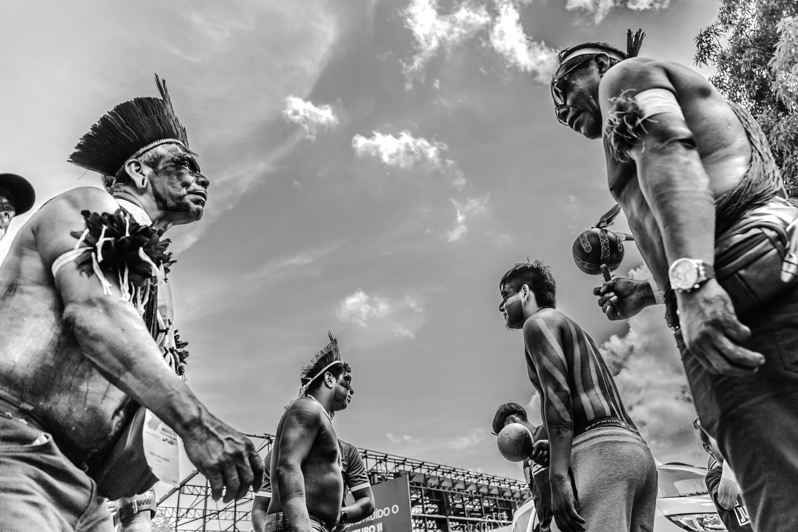 Fotografia em preto e branco mostra um grupo de homens indígenas reunidos em círculo, vistos de baixo para cima, em um momento de ritual ou manifestação cultural. Eles estão com o corpo pintado com grafismos tradicionais, usam cocares, adornos e seguram instrumentos como maracás. As expressões são sérias e concentradas, sugerindo força, ancestralidade e união. Ao fundo, há o céu com algumas nuvens e estruturas de palco, indicando que a cena acontece em um espaço público ou evento.