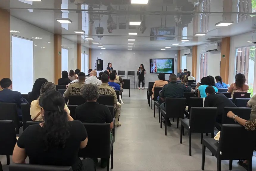 Sala de auditório com dezenas de pessoas sentadas em cadeiras pretas, voltadas para a frente. Duas mulheres estão em pé apresentando um conteúdo, ao lado de um telão com slide projetado. Parte do público veste uniformes, incluindo bombeiros militares. O ambiente é fechado, bem iluminado, com paredes claras e janelas laterais.