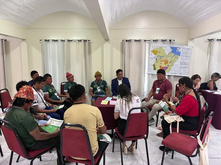 Grupo de pessoas indígenas e facilitadores sentados em círculo, em uma sala de reunião, participando de uma atividade coletiva de diálogo e leitura de materiais impressos. Ao fundo, há cortinas claras e um mapa fixado na parede, indicando um momento de discussão ou dinâmica de trabalho colaborativo durante um encontro ou seminário. Participantes utilizam crachás e vestimentas tradicionais e cotidianas.