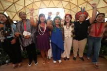 Grupo de mulheres indígenas em um espaço coberto por uma estrutura de madeira, de mãos dadas e com os punhos erguidos, em gesto de mobilização e celebração. Elas usam colares, grafismos corporais, cocares e adereços tradicionais de diferentes povos. Algumas vestem roupas modernas combinadas com elementos indígenas. Ao fundo, há outras pessoas acompanhando o momento. A cena transmite união, força coletiva, protagonismo feminino indígena e luta por direitos.