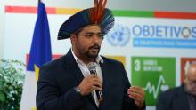 Homem adulto, de barba curta, veste terno escuro e camisa branca, segurando um microfone com a mão direita enquanto fala. Na cabeça, usa um cocar indígena colorido, com penas em tons de azul, verde e vermelho. Ele está em pé, com postura firme, e parece estar fazendo uma apresentação ou discurso.  Ao fundo, há um painel com o logotipo da ONU e a frase “Objetivos de Desenvolvimento Sustentável”, além de ícones coloridos dos ODS. À esquerda, aparece parcialmente uma bandeira azul com estrelas amarelas.