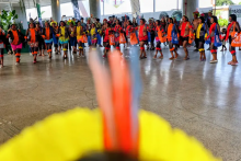 Grupo de pessoas, em sua maioria mulheres indígenas, forma uma roda e dança em um espaço coberto de piso liso. Elas vestem roupas coloridas, com destaque para tons de laranja, vermelho e amarelo, além de acessórios tradicionais. Ao fundo, há mais participantes e observadores acompanhando a atividade. Em primeiro plano, desfocado, aparece um adorno com penas coloridas, criando profundidade na imagem. O ambiente parece amplo, com pilares e iluminação natural.