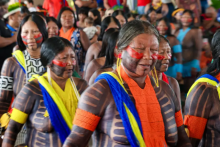 A imagem retrata um grupo de mulheres indígenas em um momento de celebração ou cerimônia, destacando-se pelas tradicionais pinturas corporais em tons de preto e vermelho. Elas utilizam adornos vibrantes, como colares e faixas de miçangas nas cores azul, amarelo e laranja, que contrastam com os grafismos na pele. A composição em plano médio foca na expressividade das mulheres à frente, enquanto o fundo preenchido por outras pessoas reforça o sentido de coletividade e resistência cultural.