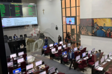 Vista de cima, a imagem mostra o plenário da Assembleia Legislativa do Pará, com deputados e assessores distribuídos em fileiras de computadores e cadeiras vermelhas. Um grande painel eletrônico à esquerda monitora as votações, enquanto um mural artístico e monitores de transmissão compõem o ambiente formal de trabalho legislativo.